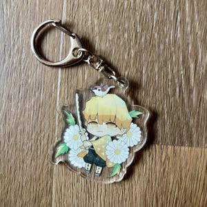 May include: A clear acrylic keychain featuring a cartoon character with yellow hair, holding a sword, and a bird on its head. The character is surrounded by white flowers and green leaves. The keychain has a silver clasp.