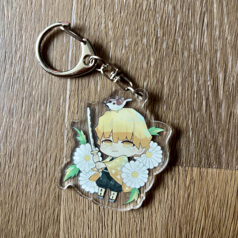 May include: A clear acrylic keychain featuring a cartoon character with yellow hair, holding a sword, and a bird on its head. The character is surrounded by white flowers and green leaves. The keychain has a silver clasp.