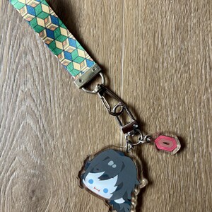 May include: A keychain featuring a cartoon character with black hair and blue eyes. The keychain has a patterned strap in green, blue, and yellow. The keychain is attached to a metal clasp and a small pink charm.