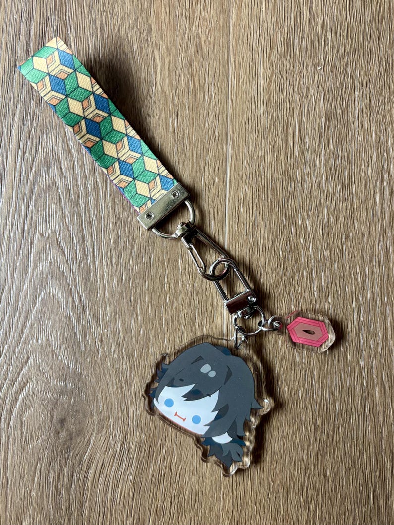 May include: A keychain featuring a cartoon character with black hair and blue eyes. The keychain has a patterned strap in green, blue, and yellow. The keychain is attached to a metal clasp and a small pink charm.