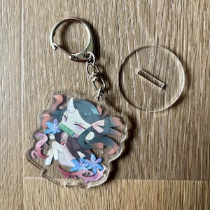 May include: A clear acrylic keychain featuring a cartoon character with dark hair, horns, and a pink bow. The character is holding a bamboo tube and surrounded by blue flowers and pink accents. A silver keyring and a clear stand are also included.