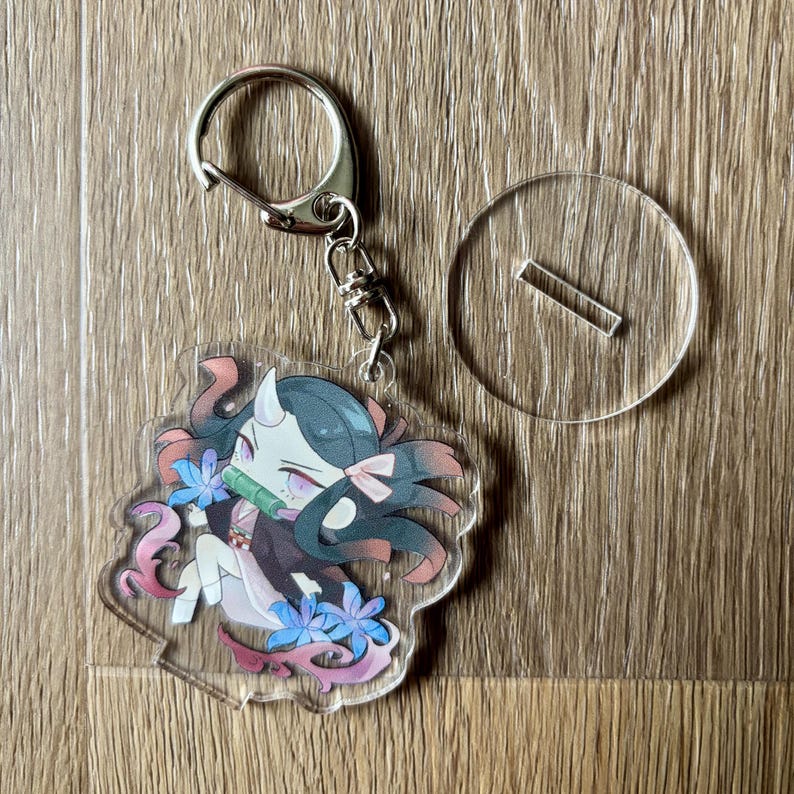 May include: A clear acrylic keychain featuring a cartoon character with dark hair, horns, and a pink bow. The character is holding a bamboo tube and surrounded by blue flowers and pink accents. A silver keyring and a clear stand are also included.