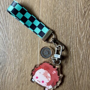 May include: A keychain featuring a cartoon character with red hair and a checkered strap in black and teal. The keychain includes a small, circular charm and a silver-colored clasp.