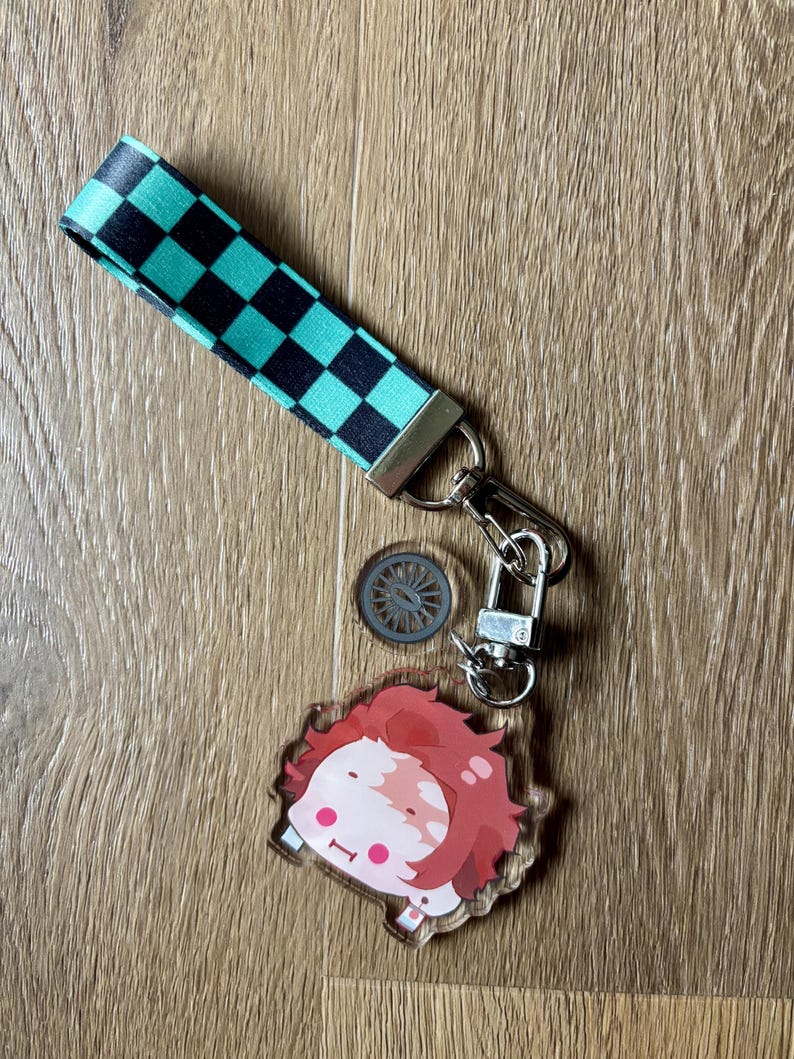 May include: A keychain featuring a cartoon character with red hair and a checkered strap in black and teal. The keychain includes a small, circular charm and a silver-colored clasp.