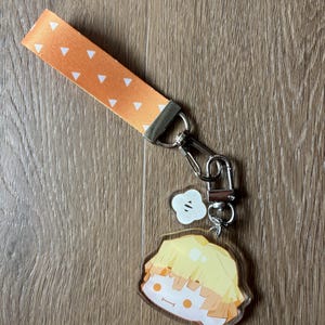 May include: An orange wrist strap with white triangle pattern, connected to a silver clasp and a charm. The charm features a cartoon character with yellow hair and a smiling face. A small white charm is also attached.
