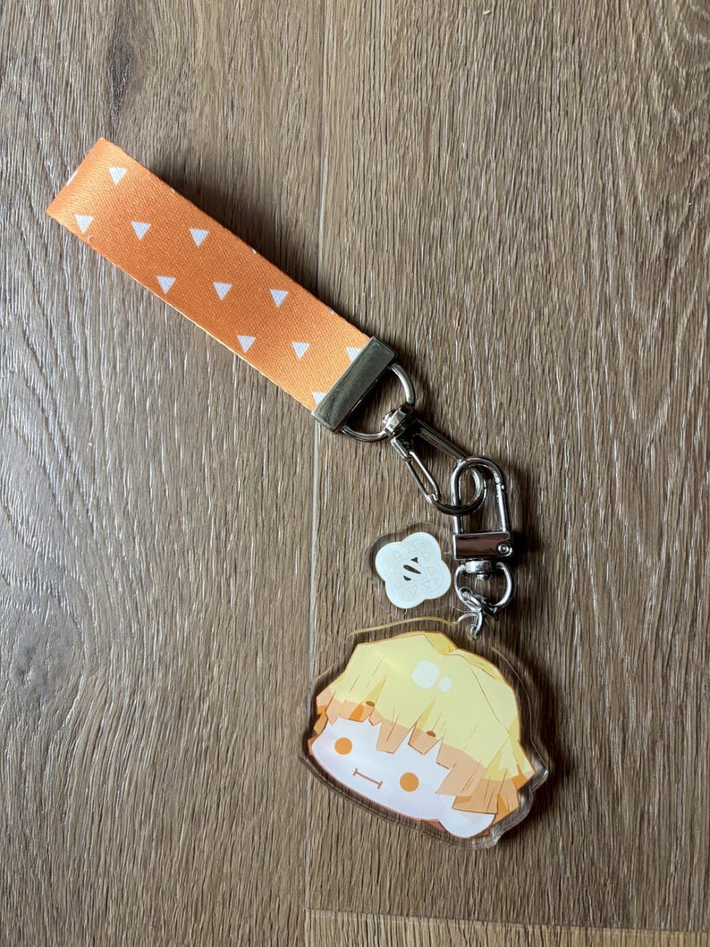 May include: An orange wrist strap with white triangle pattern, connected to a silver clasp and a charm. The charm features a cartoon character with yellow hair and a smiling face. A small white charm is also attached.