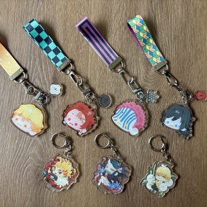 May include: A collection of character keychains with matching wrist straps. The keychains feature colorful anime-style illustrations. The wrist straps have various patterns, including checkered, striped, and geometric designs. The keychains are attached to silver-colored keyrings.