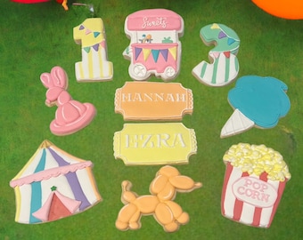 May include: A set of nine colorful decorated sugar cookies featuring a circus theme. The cookies include a pink bunny, a blue cotton candy, a red and white striped popcorn bucket, a yellow tent, a yellow and white number one, a green and white number three, a pink and white sweets cart, and two yellow tickets with the names "Hannah" and "Ezra".