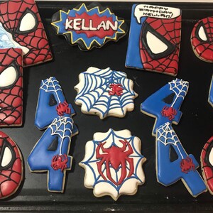 May include: A set of decorated sugar cookies featuring a Spiderman theme. The cookies are decorated with red, blue, and white icing. The cookies include a Spiderman head, a web design, and the number 4. The cookies are arranged on a black baking sheet. One cookie has the text "Happy Birthday Kellan!" on it.