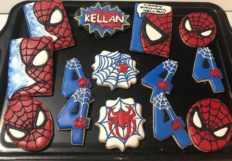 May include: A set of decorated sugar cookies featuring a Spiderman theme. The cookies are decorated with red, blue, and white icing. The cookies include a Spiderman head, a web design, and the number 4. The cookies are arranged on a black baking sheet. One cookie has the text "Happy Birthday Kellan!" on it.