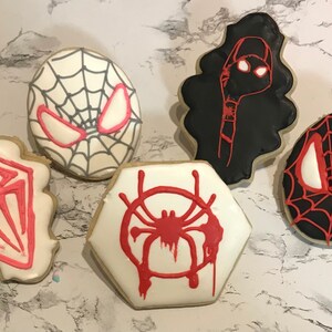 May include: Five decorated sugar cookies featuring different Spider-Man characters and symbols. The cookies are white, black, and red with black and white web designs, red spider symbols, and red and white eyes.