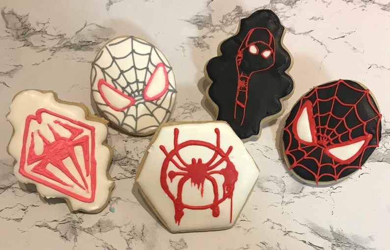 May include: Five decorated sugar cookies featuring different Spider-Man characters and symbols. The cookies are white, black, and red with black and white web designs, red spider symbols, and red and white eyes.