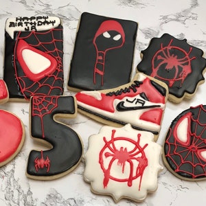 May include: A set of nine cookies decorated with a Spider-Man theme. The cookies are black, red, and white and feature images of Spider-Man, a spider web, and the number "5". One cookie has the text "Happy Birthday JA".