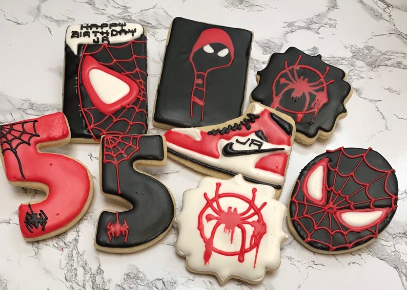 May include: A set of nine cookies decorated with a Spider-Man theme. The cookies are black, red, and white and feature images of Spider-Man, a spider web, and the number "5". One cookie has the text "Happy Birthday JA".