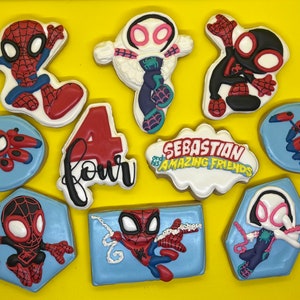 May include: A set of ten decorated sugar cookies featuring Spider-Man characters. The cookies are decorated with red, blue, black, white, and pink icing. The cookies are arranged on a yellow surface. The cookies feature the text "Sebastian's Amazing Friends" and "Four".