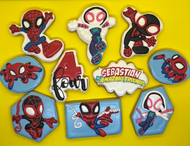 May include: A set of ten decorated sugar cookies featuring Spider-Man characters. The cookies are decorated with red, blue, black, white, and pink icing. The cookies are arranged on a yellow surface. The cookies feature the text "Sebastian's Amazing Friends" and "Four".