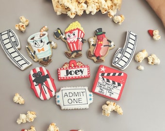May include: A set of nine movie-themed sugar cookies decorated with red, white, and blue frosting. The cookies feature images of popcorn, a film reel, a movie ticket, a clapperboard, a hot dog, a soda, and a sign that says "Joey".