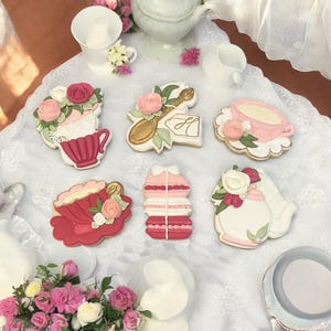 May include: Six decorated sugar cookies on a white lace tablecloth. The cookies are decorated with pink and white frosting and feature teacups, a teapot, a spoon, and macarons.