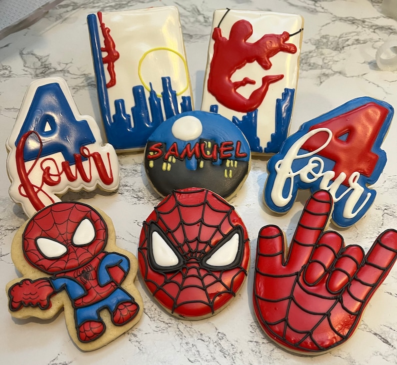 May include: A set of ten decorated sugar cookies featuring a Spiderman theme. The cookies are decorated with red, blue, and white icing. The cookies feature the number four, the words "four", a Spiderman mask, and a Spiderman hand.