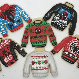 May include: Six decorated sugar cookies shaped like sweaters with a Spider-Man theme. The sweaters are red, blue, black, green, and white with various patterns and Spider-Man imagery.