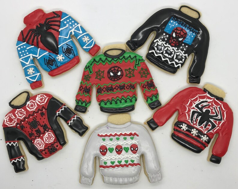 May include: Six decorated sugar cookies shaped like sweaters with a Spider-Man theme. The sweaters are red, blue, black, green, and white with various patterns and Spider-Man imagery.