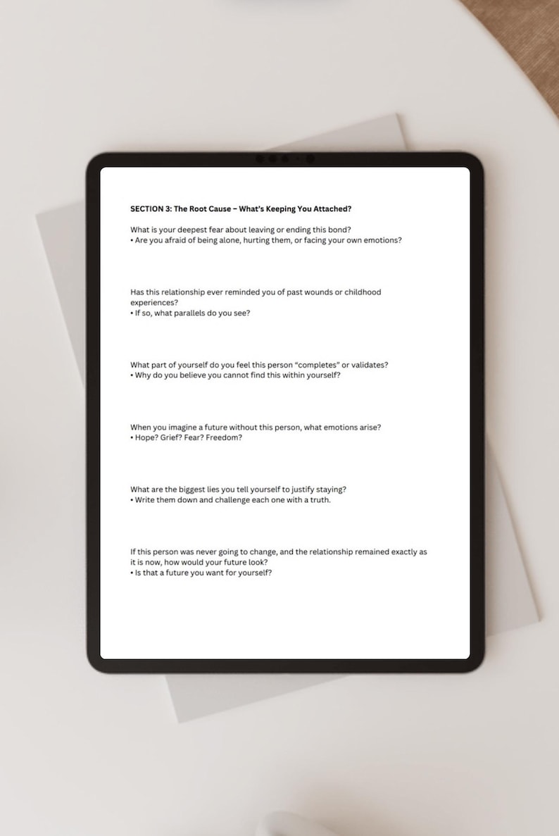 May include: A black tablet displaying text from "SECTION 3: The Root Cause - What's Keeping You Attached?" The text includes questions about relationships, fears, and self-reflection. The tablet is on a white surface.