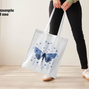 White tote bag with a blue moth and floral design. The moth has a dark blue body and light blue wings. The floral design is in shades of blue and white.