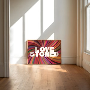 May include: A framed art print with the words "LOVE STONED" in white block letters. The artwork features a colorful, swirling pattern of red, pink, orange, green, and purple. The frame is a light brown color, and the print is displayed on a wooden floor.