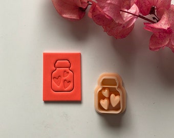 Puede incluir: Dos cortadores de galletas, uno rojo y otro melocotón, con forma de tarro con corazones en su interior.