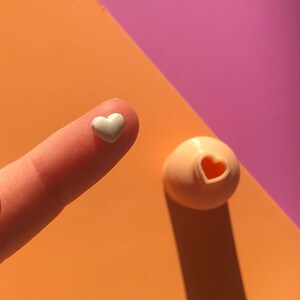 May include: A white heart-shaped object on a finger, next to a pink heart-shaped mold on a pink and orange surface.