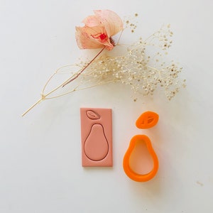 Könnte beinhalten: Zwei Ausstecher aus rosa und orangefarbenem Polymerton in Form von Birnen. Die Ausstecher liegen auf einer weißen Oberfläche mit einem Trockenblumenarrangement im Hintergrund.