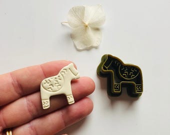 May include: A white and a green plastic cookie cutter in the shape of a traditional Swedish Dala horse. The white cookie cutter is being held in a hand.