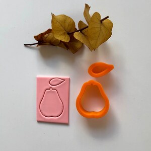 Könnte beinhalten: Ein Set aus zwei orangefarbenen Keksausstechern in Form von Birnen. Ein Ausstecher ist durchgehend, der andere ist eine Umrandung. Eine rosa Plastikform einer Birne mit einem Blatt ist ebenfalls enthalten.