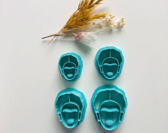 May include: Four blue plastic cookie cutters shaped like a person's head with a face and hair. The cutters are on a white surface.