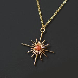 K&ouml;nnte beinhalten: Eine goldfarbene Halskette mit einem Anh&auml;nger in Form eines Sonnenstrahls, der einen orange-roten Stein in der Mitte aufweist. Der Anh&auml;nger hat strahlenf&ouml;rmige Linien, und die Kette ist filigran. Der Schmuck wird vor einem dunklen Hintergrund pr&auml;sentiert.