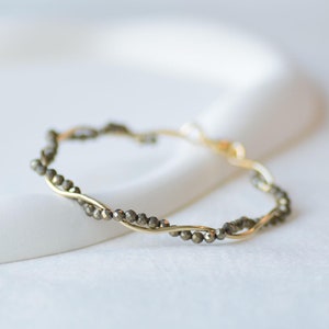 May include: A gold and pyrite beaded bracelet with a twisted design. The bracelet is on a white surface.