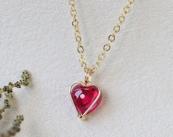 May include: A gold-colored chain necklace with a heart-shaped pendant. The pendant is a vibrant red gemstone encased in a gold wire frame. The necklace is displayed on a white surface with a sprig of greenery.