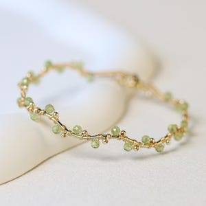 May include: A delicate gold wire ring featuring a series of small, faceted light green peridot gemstones spaced evenly along the band. The wire is twisted into a subtle, organic vine-like pattern, securing each individual stone in place.