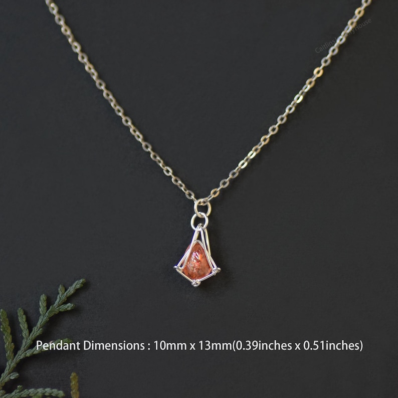 May include: A silver-toned chain necklace featuring a small, teardrop-shaped orange gemstone pendant encased in a silver wire cage. The pendant measures 0.39 inches by 0.51 inches. The necklace is displayed against a dark, solid background.