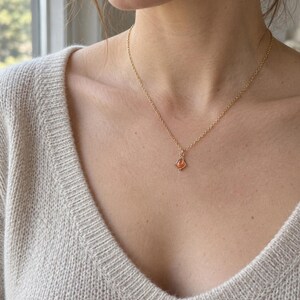 May include: A delicate gold chain necklace featuring a small, teardrop-shaped orange gemstone pendant encased in a thin gold wire frame. The necklace rests against a beige knit sweater with a V-neckline, highlighting the minimalist jewelry design.