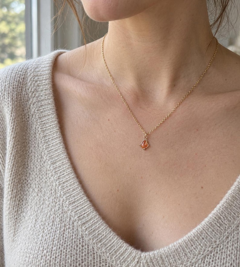 May include: A delicate gold chain necklace featuring a small, teardrop-shaped orange gemstone pendant encased in a thin gold wire frame. The necklace rests against a beige knit sweater with a V-neckline, highlighting the minimalist jewelry design.