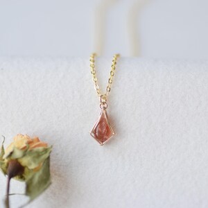 May include: A gold chain necklace featuring a kite-shaped pendant with a translucent, reddish-orange gemstone. The pendant is held by a delicate gold wire frame, and the chain is thin with small, uniform oval links, creating a simple, elegant piece.