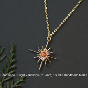 K&ouml;nnte beinhalten: Eine goldfarbene Halskette mit einem Anh&auml;nger in Form eines Sonnenstrahls. Der Anh&auml;nger hat einen zentralen orange-roten Stein und strahlenf&ouml;rmige Speichen aus Draht. Die Kette ist filigran, und der Hintergrund ist dunkel. Der Text "Handmade &bull; Slight Variations (&plusmn;1-2mm) &bull; Subtle Handmade Marks" steht unten.