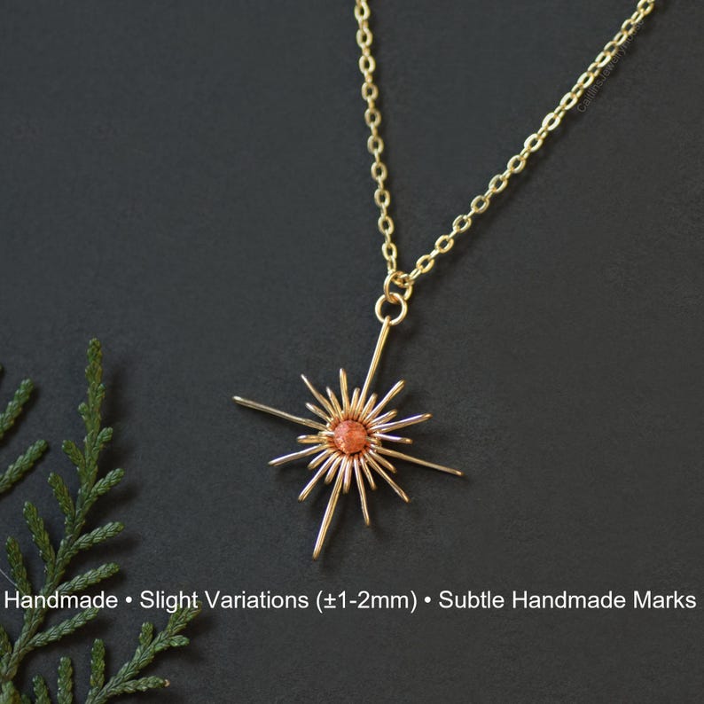 K&ouml;nnte beinhalten: Eine goldfarbene Halskette mit einem Anh&auml;nger in Form eines Sonnenstrahls. Der Anh&auml;nger hat einen zentralen orange-roten Stein und strahlenf&ouml;rmige Speichen aus Draht. Die Kette ist filigran, und der Hintergrund ist dunkel. Der Text "Handmade &bull; Slight Variations (&plusmn;1-2mm) &bull; Subtle Handmade Marks" steht unten.