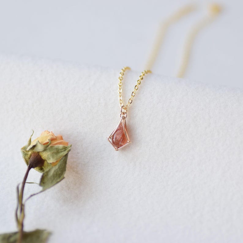 May include: A delicate gold chain necklace featuring a small, kite-shaped pendant with a translucent, reddish-orange gemstone set in a thin gold frame. The necklace is positioned near a dried, pale orange rose with green leaves.