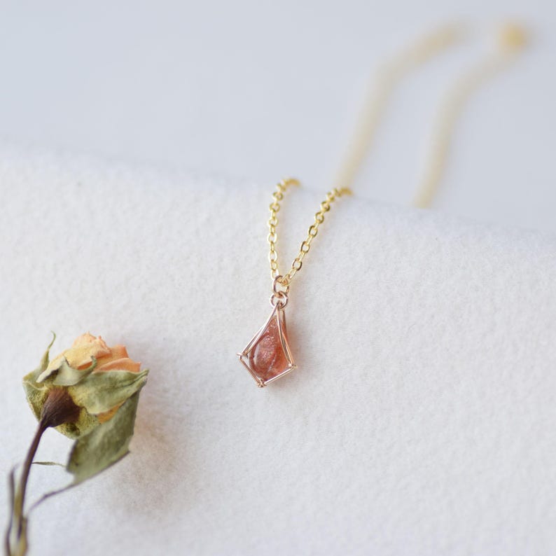 May include: A gold chain necklace with a pendant featuring a small, reddish-brown gemstone encased in a gold wire cage.