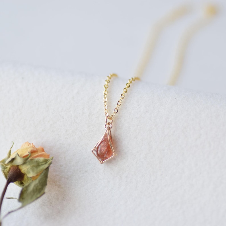 May include: A delicate gold chain necklace featuring a small, kite-shaped pendant. The pendant consists of a polished, translucent orange-red gemstone encased within a thin, geometric gold wire frame that mirrors the kite shape.