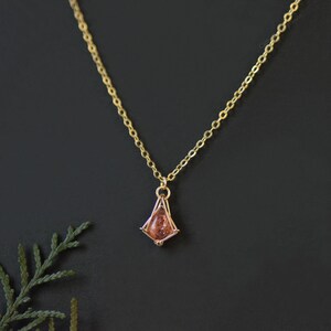 May include: A gold chain necklace featuring a small, kite-shaped pendant. The pendant consists of a metallic frame holding a single, translucent, peach-colored gemstone with visible internal flecks and a teardrop-like shape.