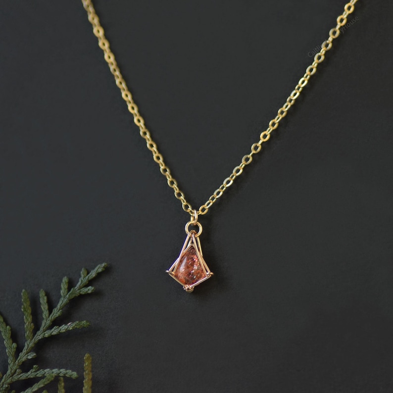 May include: A gold chain necklace featuring a small, kite-shaped pendant. The pendant consists of a metallic frame holding a single, translucent, peach-colored gemstone with visible internal flecks and a teardrop-like shape.