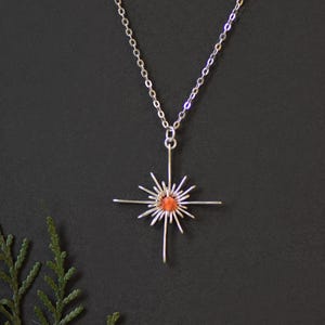 K&ouml;nnte beinhalten: Eine silberfarbene Halskette mit Anh&auml;nger, die ein Sonnenstrahlen-Design aufweist. Der Anh&auml;nger hat einen zentralen orange-roten Stein, der von strahlenden silbernen Linien umgeben ist. Die Halskette h&auml;ngt an einer filigranen Silberkette vor dunklem Hintergrund.